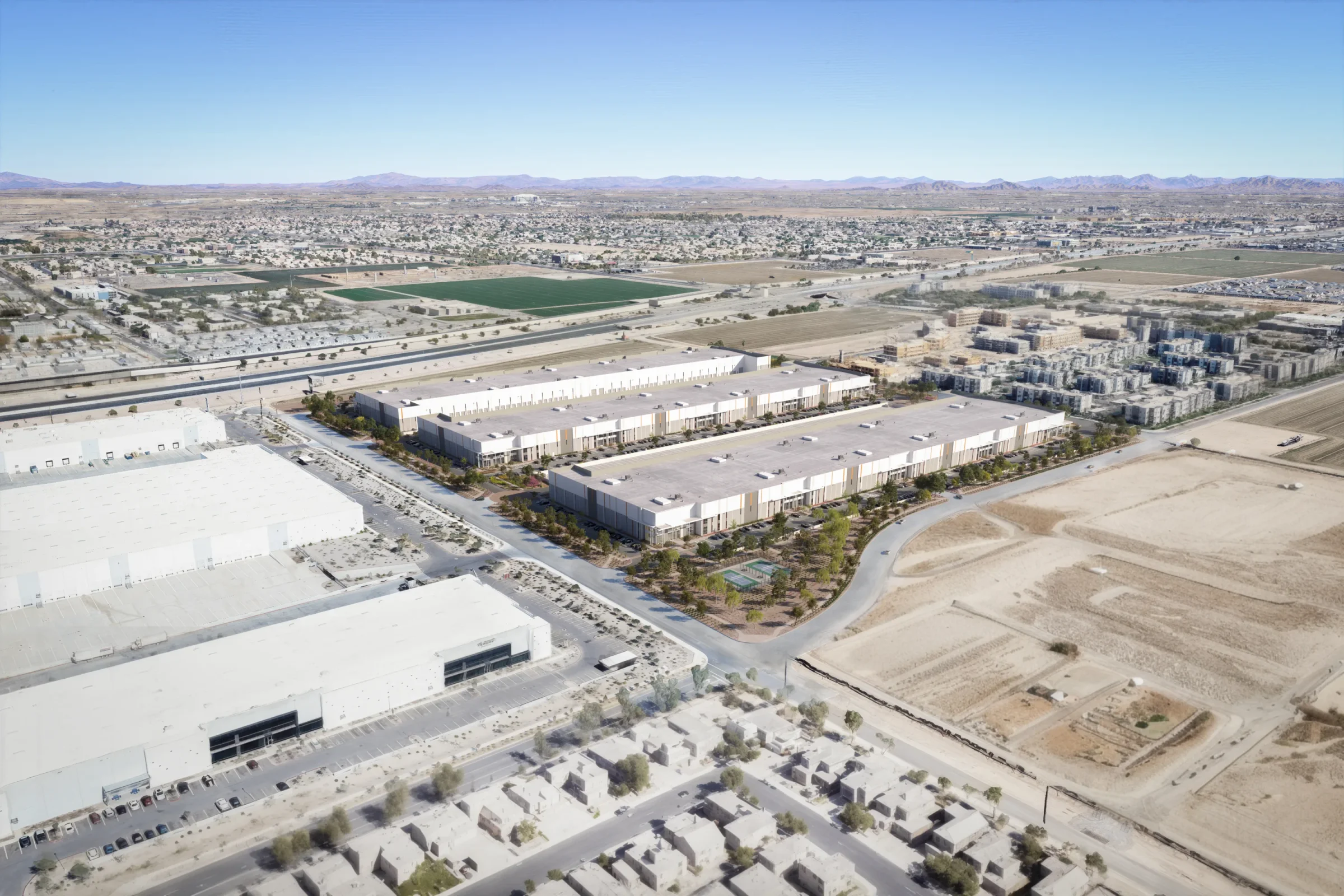 Aerial view of a light industrial/business park development with several long, low-rise white warehouse or flex-space buildings arranged in rows, surrounded by landscaped walkways and parking lots. In the foreground are additional large white warehouse rooftops. The background shows a residential neighborhood, agricultural fields, open desert land, and a mountain range under a clear blue sky.