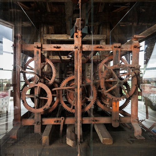 Complex arrangement of large, rusty gears and mechanical parts encased in glass, situated in a wooden structure.