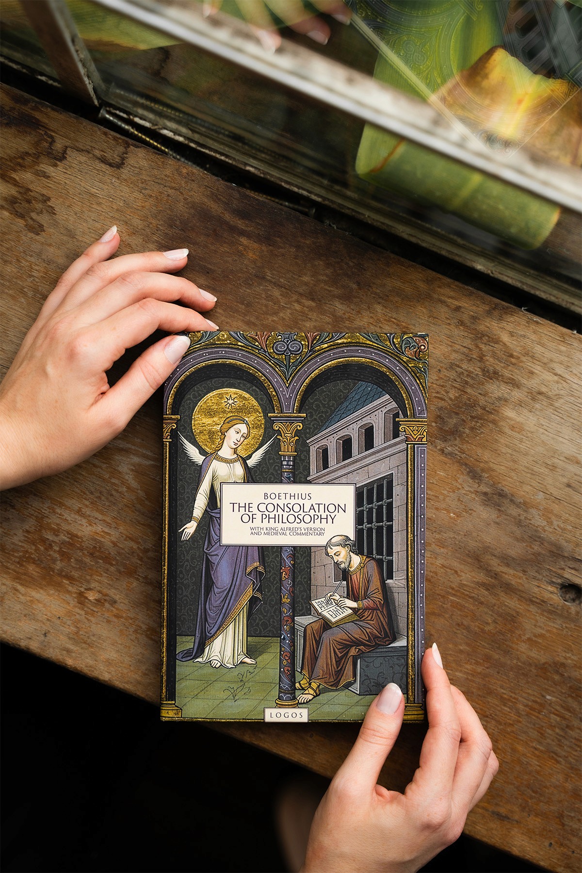 Hands holding the book The Consolation of Philosophy by Boethius, published by Logos, on a rustic wooden surface.