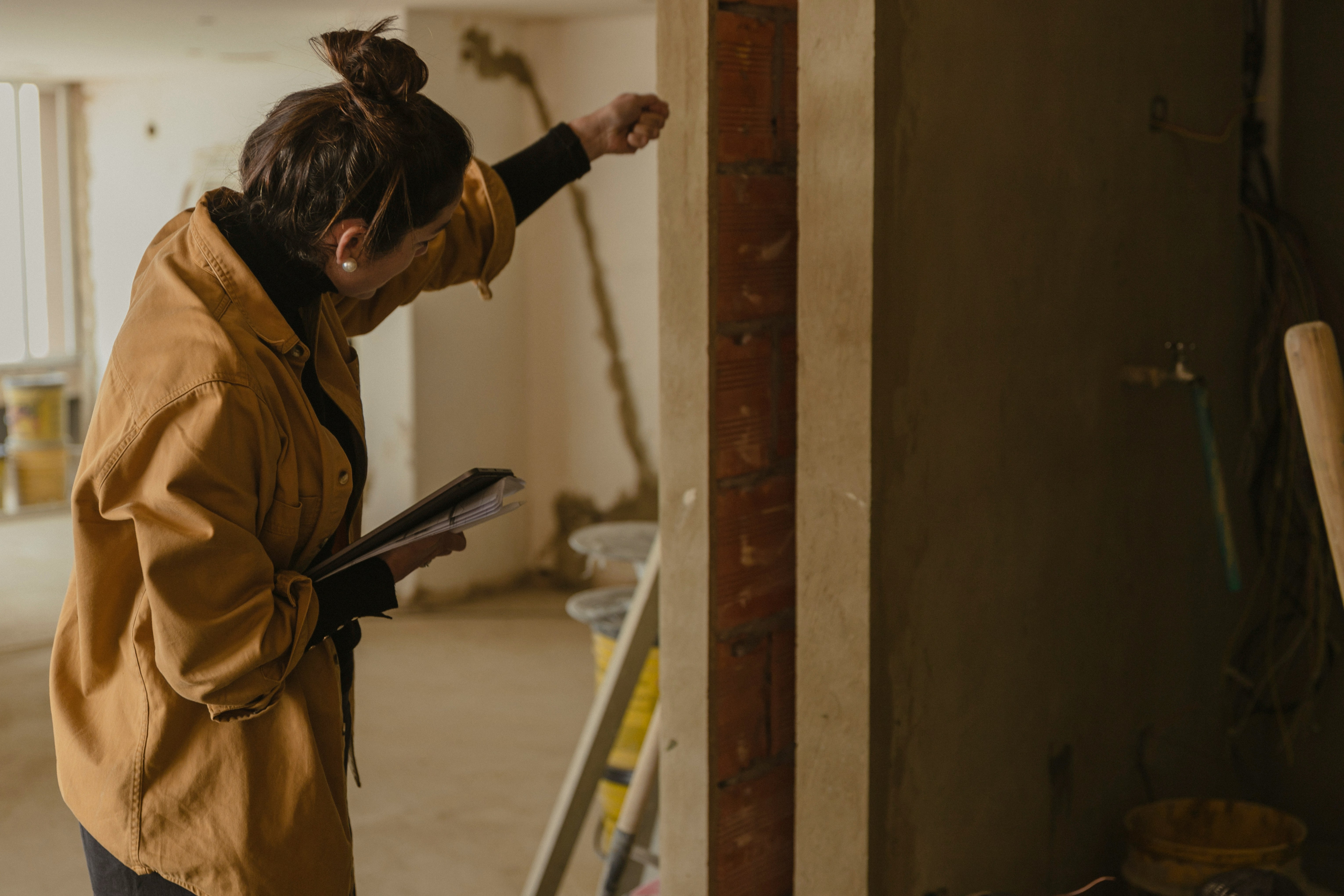 A person in a brown jacket stands near a ladder, working on a wall in an unfinished room.