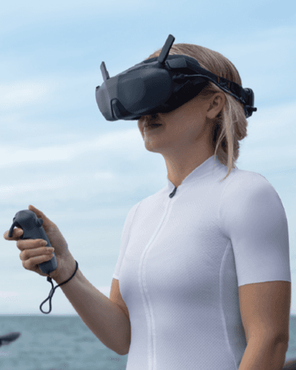 A person wearing virtual reality goggles and holding a controller, with a scenic background of water and sky.