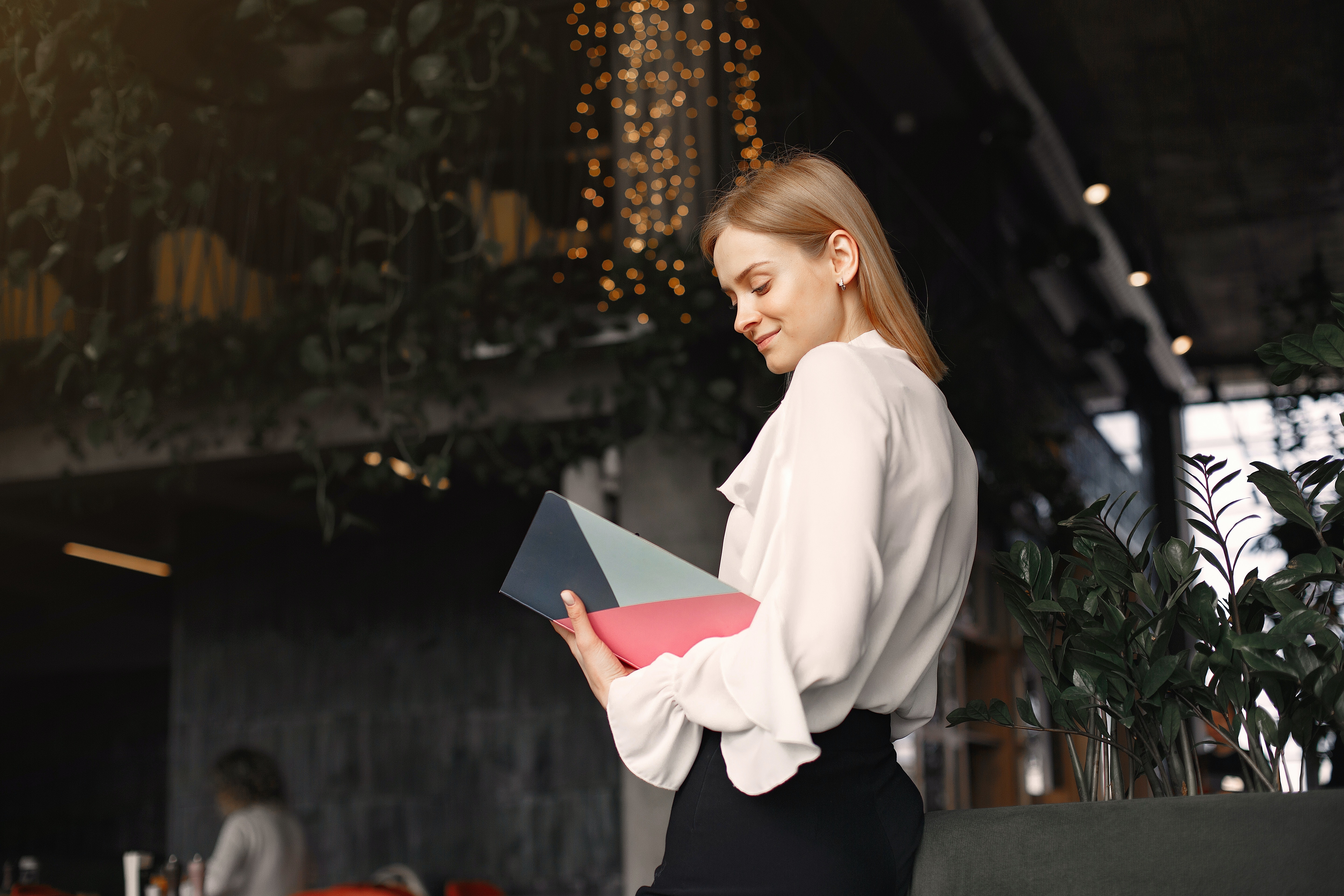 Een jonge vrouw in een witte blouse staat buiten, met een tijdschrift in haar hand en kijkt ernaar.
