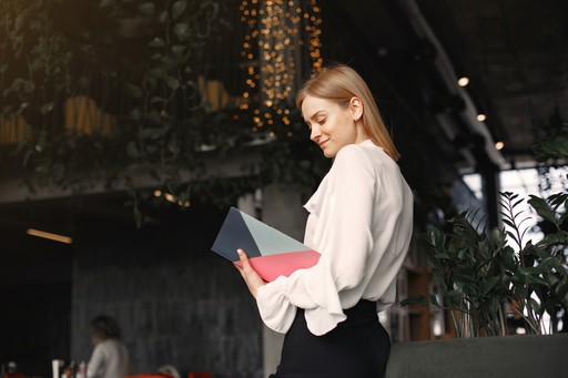 A young woman in a white blouse stands outdoors, holding a magazine and looking down at it.