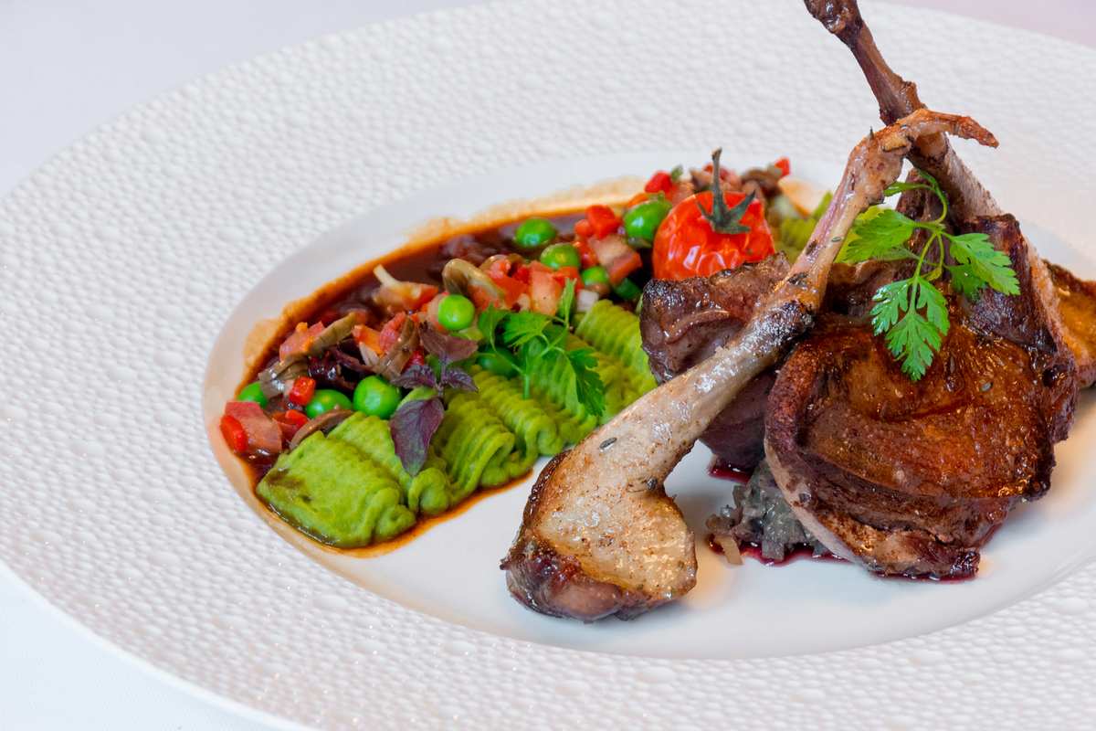 A plated dish featuring roasted duck leg and breast served with avocado slices, cherry tomatoes, and fresh herbs on a white