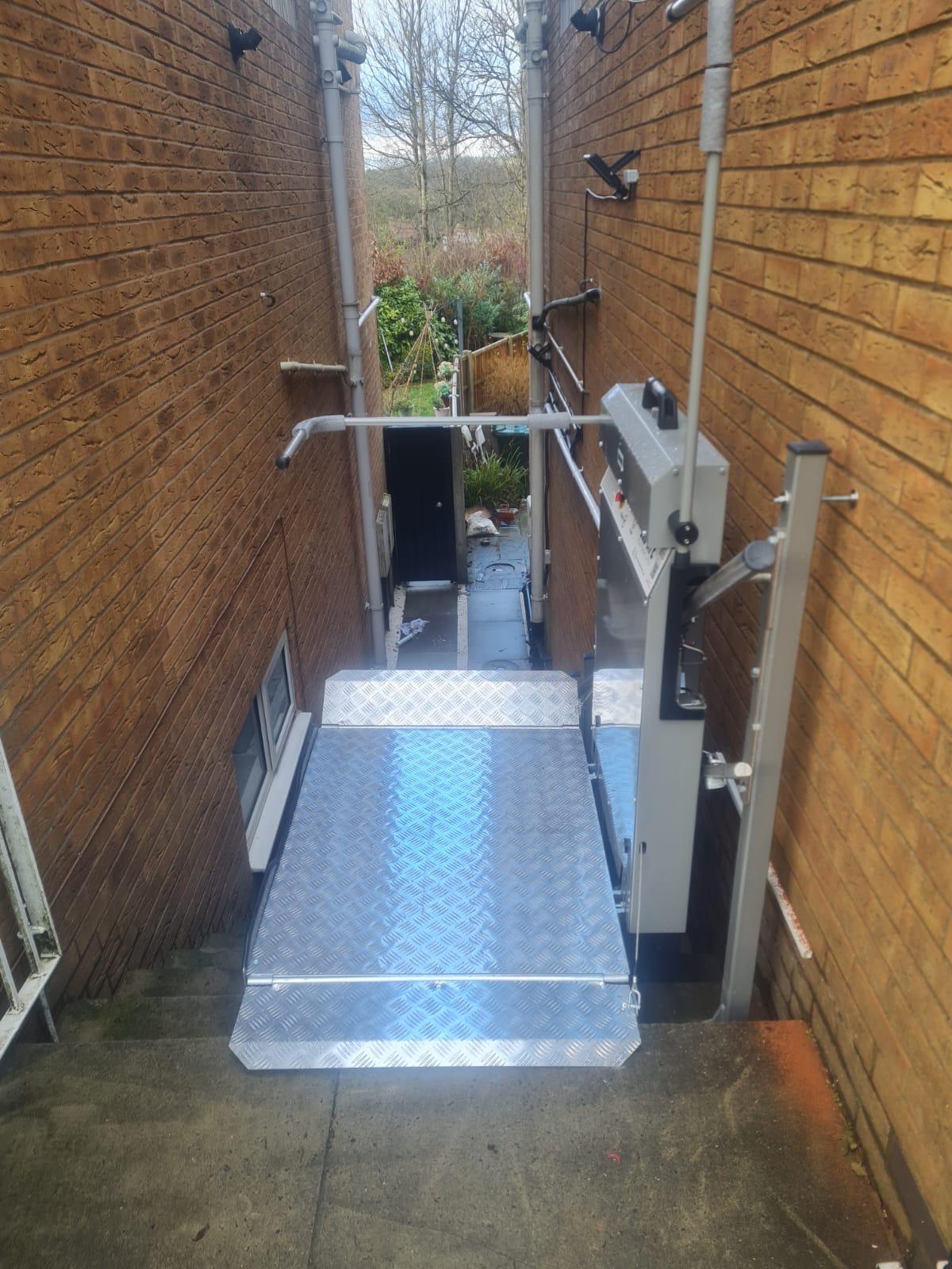 Vertical platform lift installed in a narrow external passageway between two brick walls at a UK residential property — anti-slip chequer plate platform at lower landing level with drive column and handrails visible