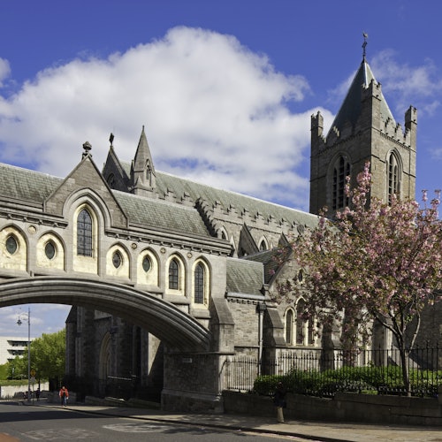Gothic-style stone cathedral with an arched bridge, a flowering tree, and clear blue sky. Two people walking below.