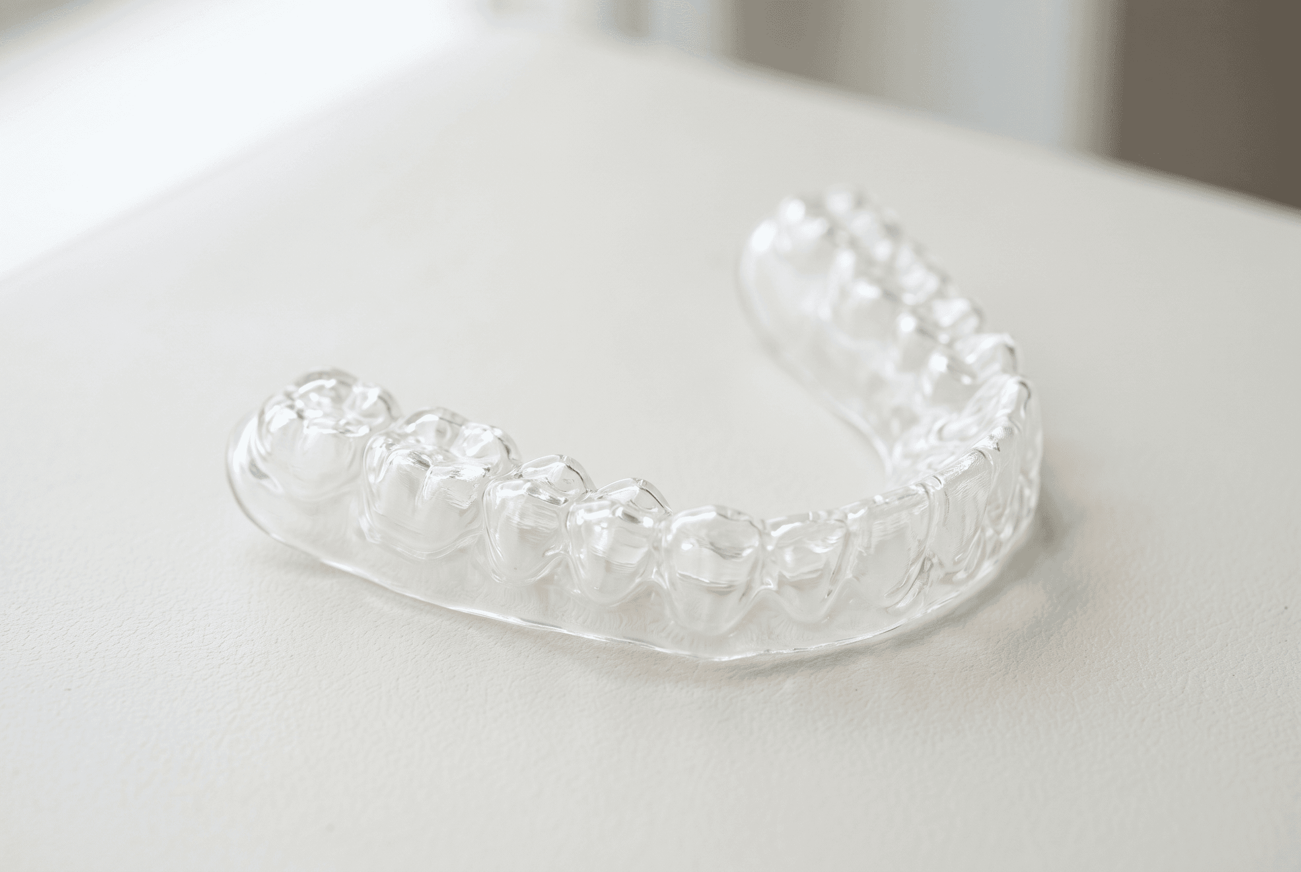 Macro of a clear transparent dental splint resting on a pale surface, catching soft daylight along its edges, clean and precise, medical device product photography