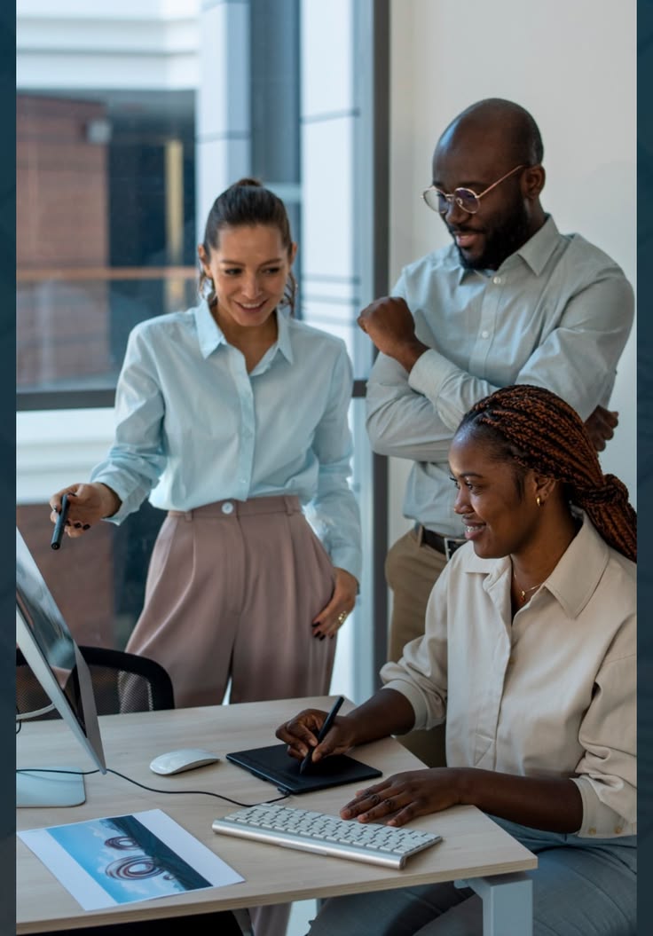 A man wearing glasses and a blazer working at a desk, writing in a notebook beside a computer monitor in a modern office.