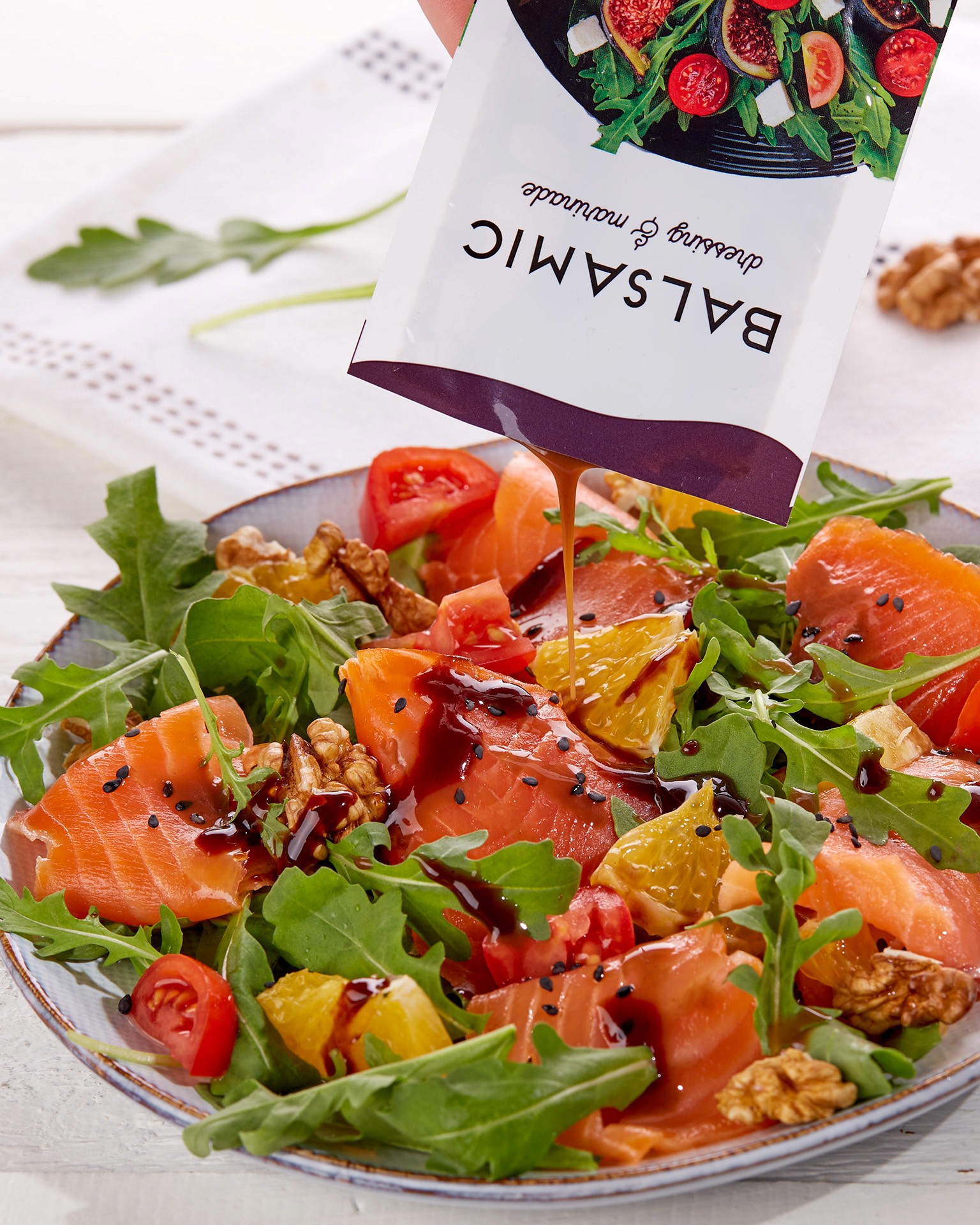 A fresh salad featuring arugula, sliced tomatoes, smoked salmon, orange segments, and walnuts is being drizzled with balsamic dressing from a packet.