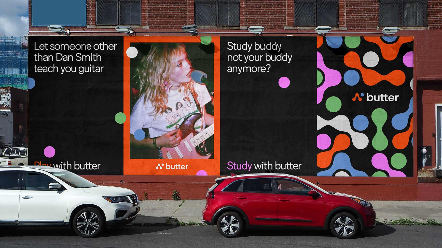 A brick building displays three large billboards. The leftmost billboard reads “Let someone other than Dan Smith teach you guitar.” The center billboard shows a blonde girl playing guitar. The rightmost billboard displays a black background with colorful abstract shapes and the word “butter” in white text. Two cars are parked on the street in front of the billboards.