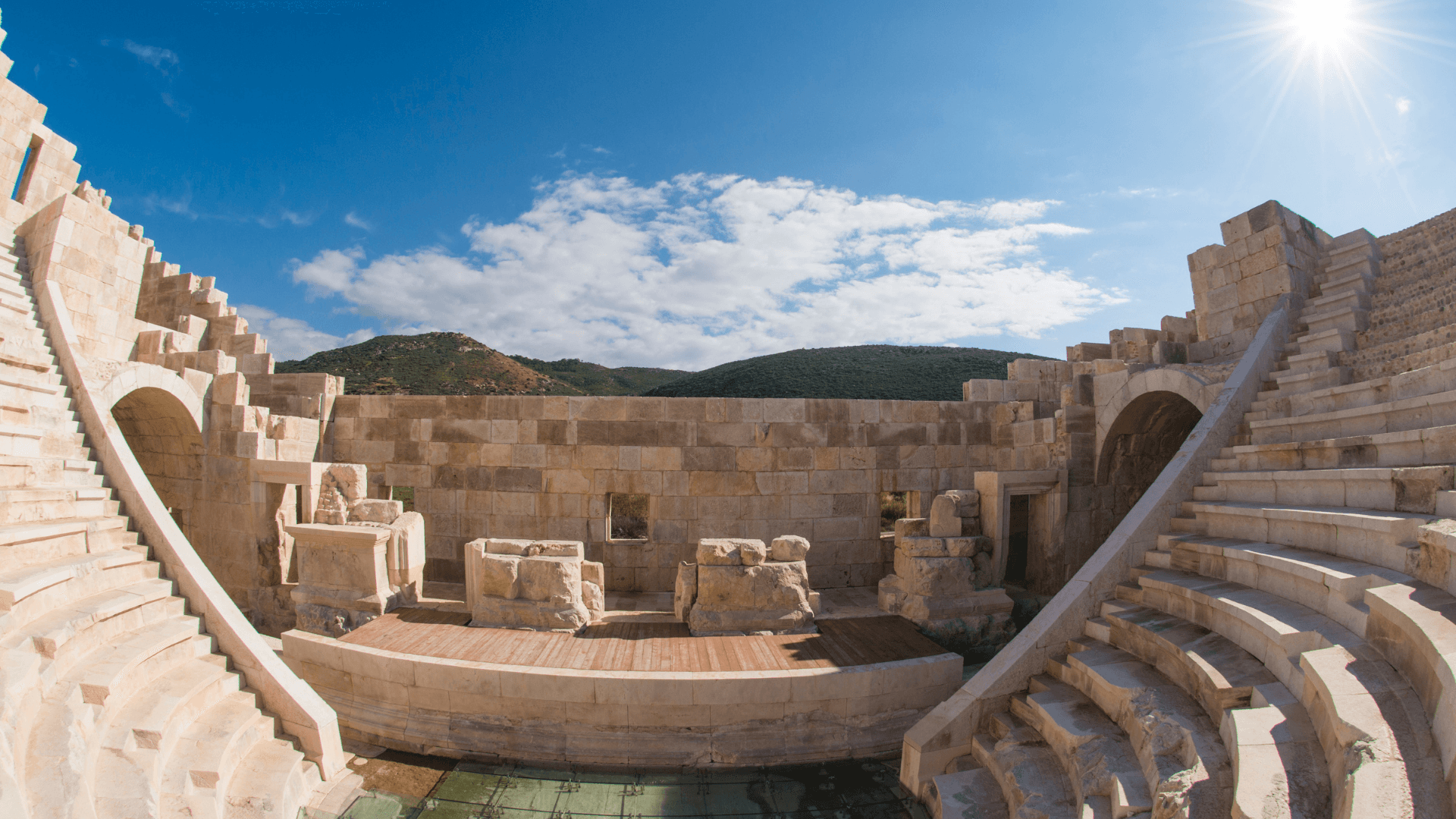 Ancient Roman theater ruins in Patara, Türkiye — Restored stone seating and stage with scenic hills and a bright sun overhead.
