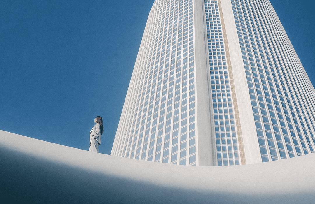 Mulher de terno observando um arranha-céu moderno sob céu azul claro. Estilo visual minimalista e futurista da Inspira.