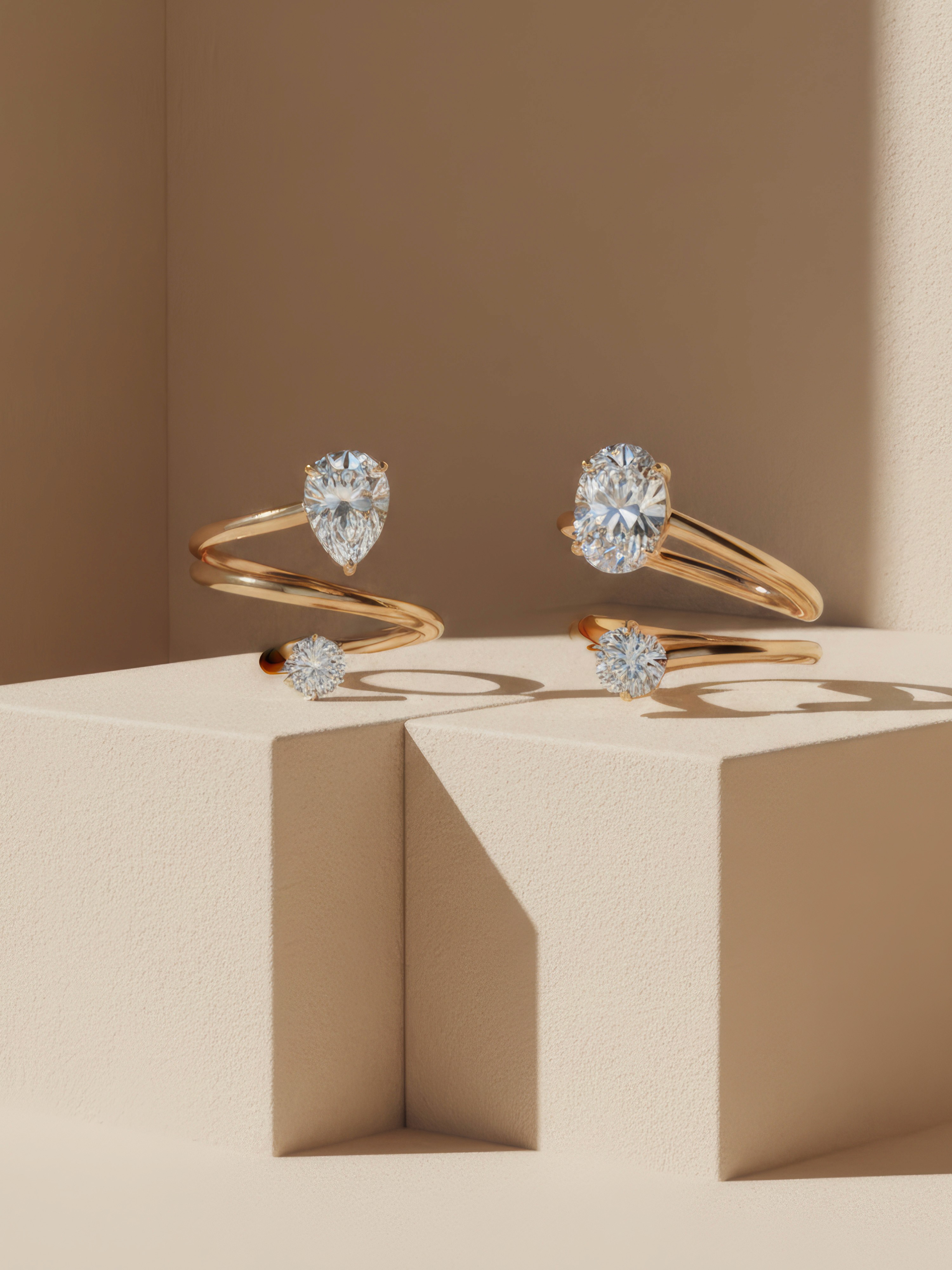 Render fotorealistico di gioielleria: coppia di anelli a spirale in oro giallo con diamanti solitari (taglio a goccia e taglio ovale). Allestimento su podi geometrici color beige con illuminazione solare e ombre nette.