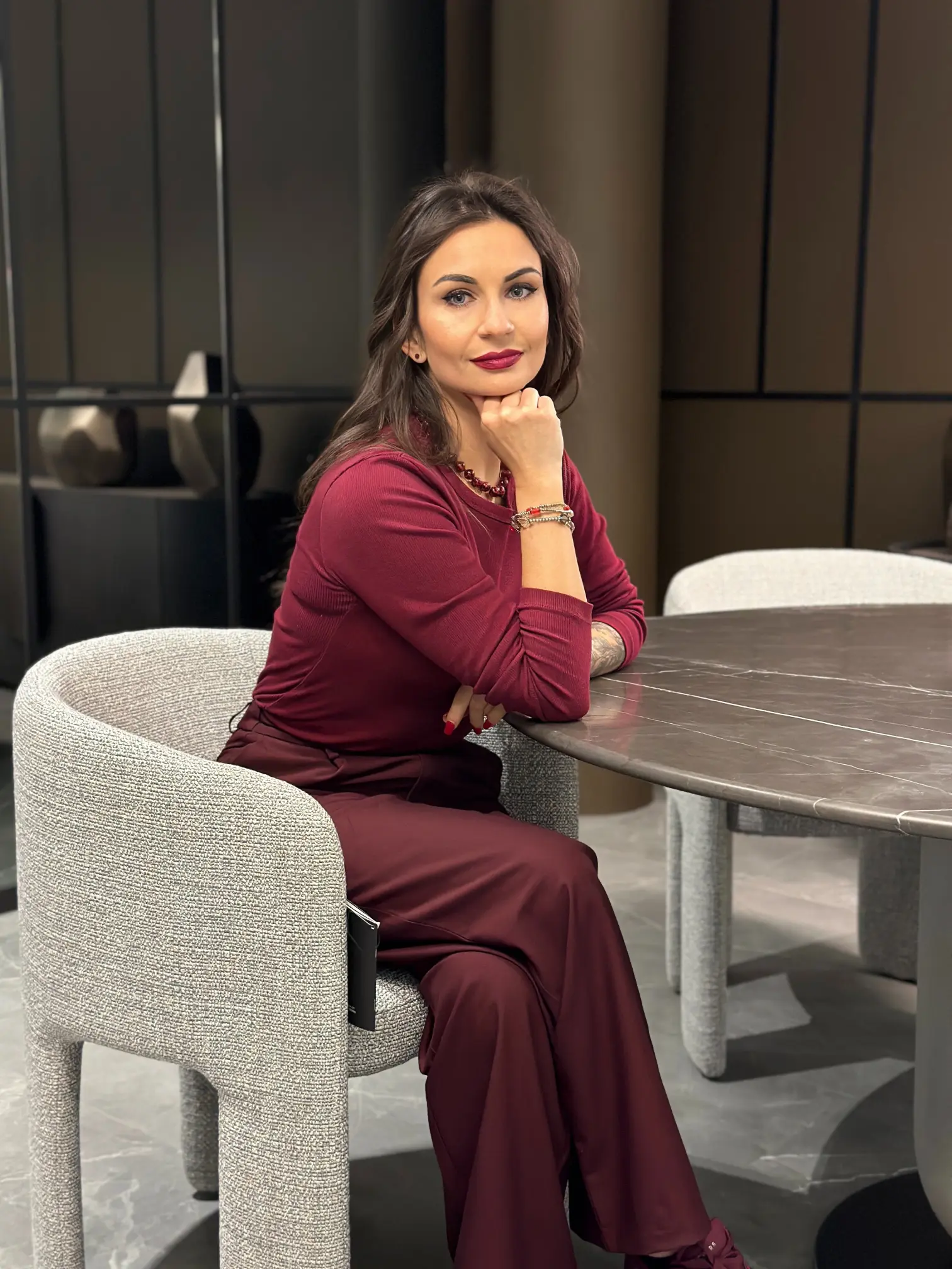 Projektantka w bordowej sukience siedzi na kanapie w otoczeniu roślin.