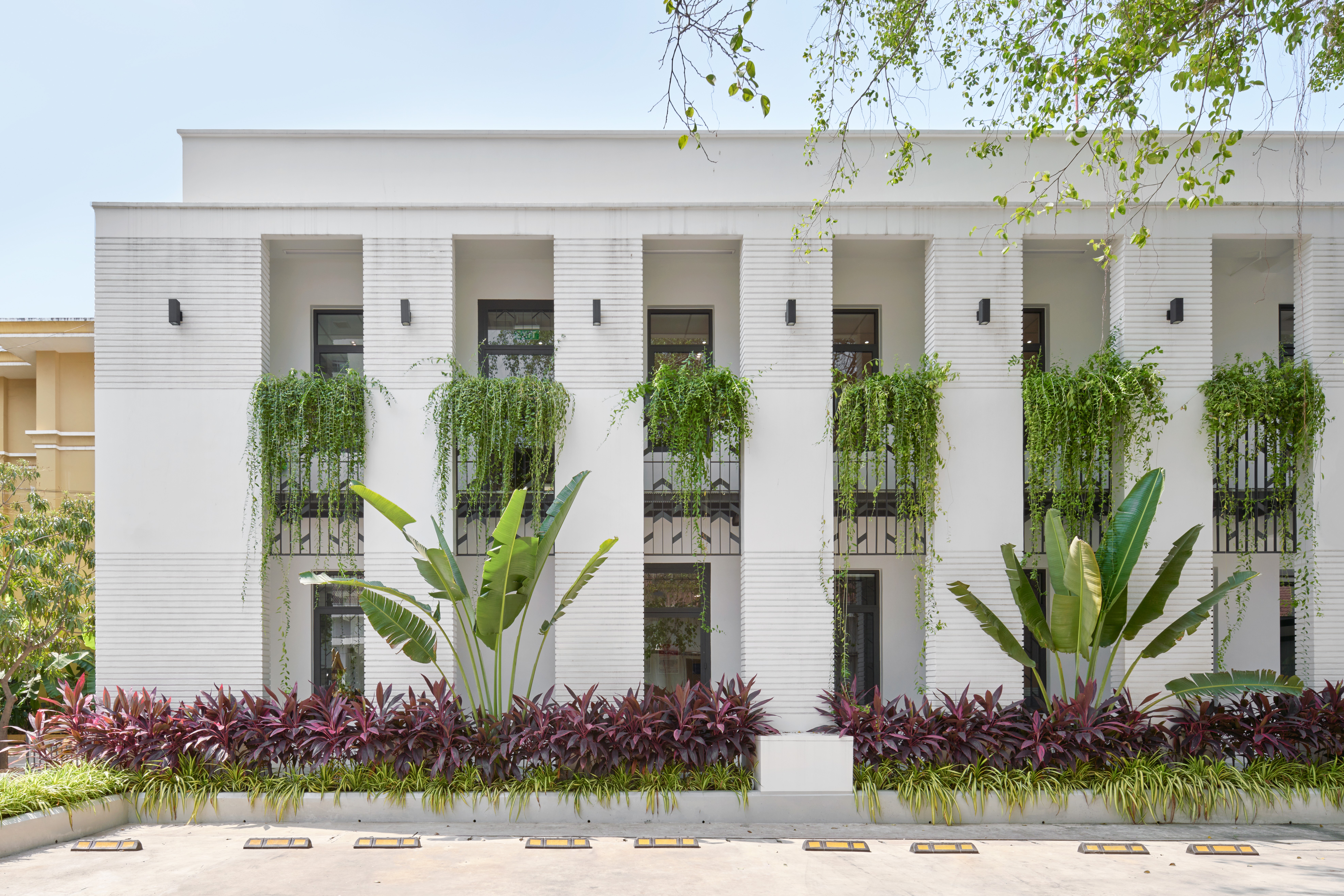 Flat architectural elevation of a white office building showing repetitive vertical bays, black window frames, and vibrant tropical landscaping.