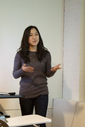 A photo of a woman standing near a classroom table and giving a lecture.