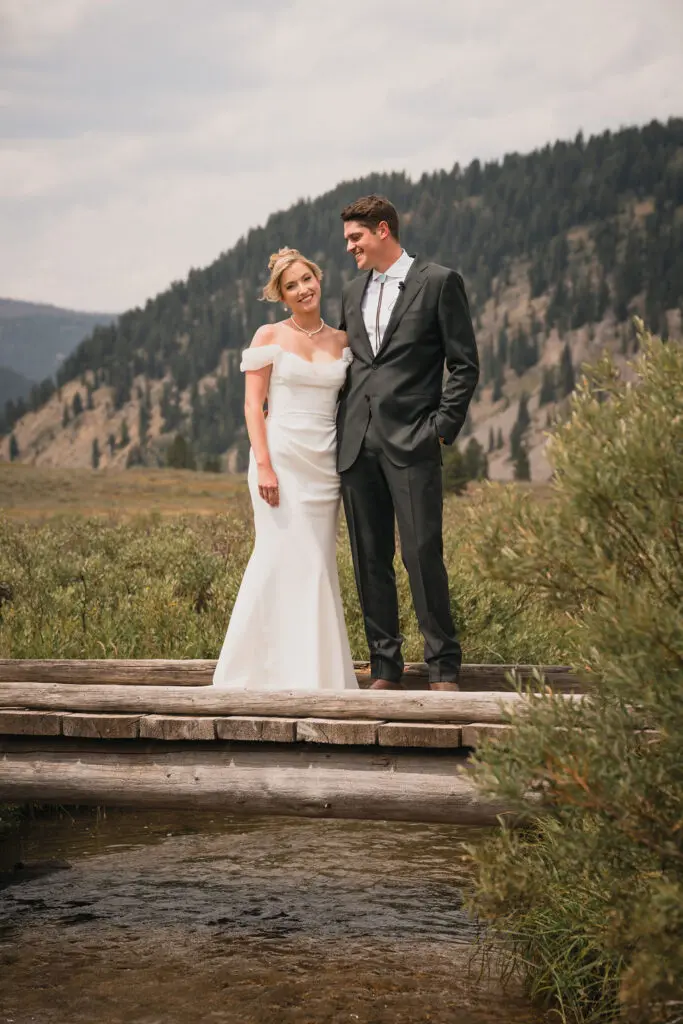 The bride and groom pose together on a rustic log bridge over a small creek, with a backdrop of a forested mountain and a cloudy sky.