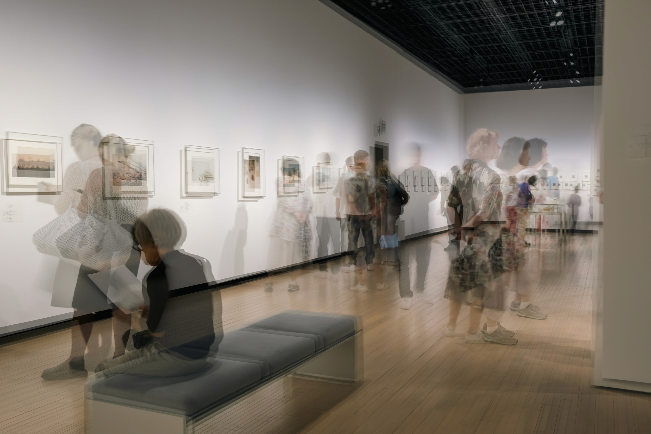 Black and white slow shutter photograph showing blurred figures walking through a contemporary art gallery.