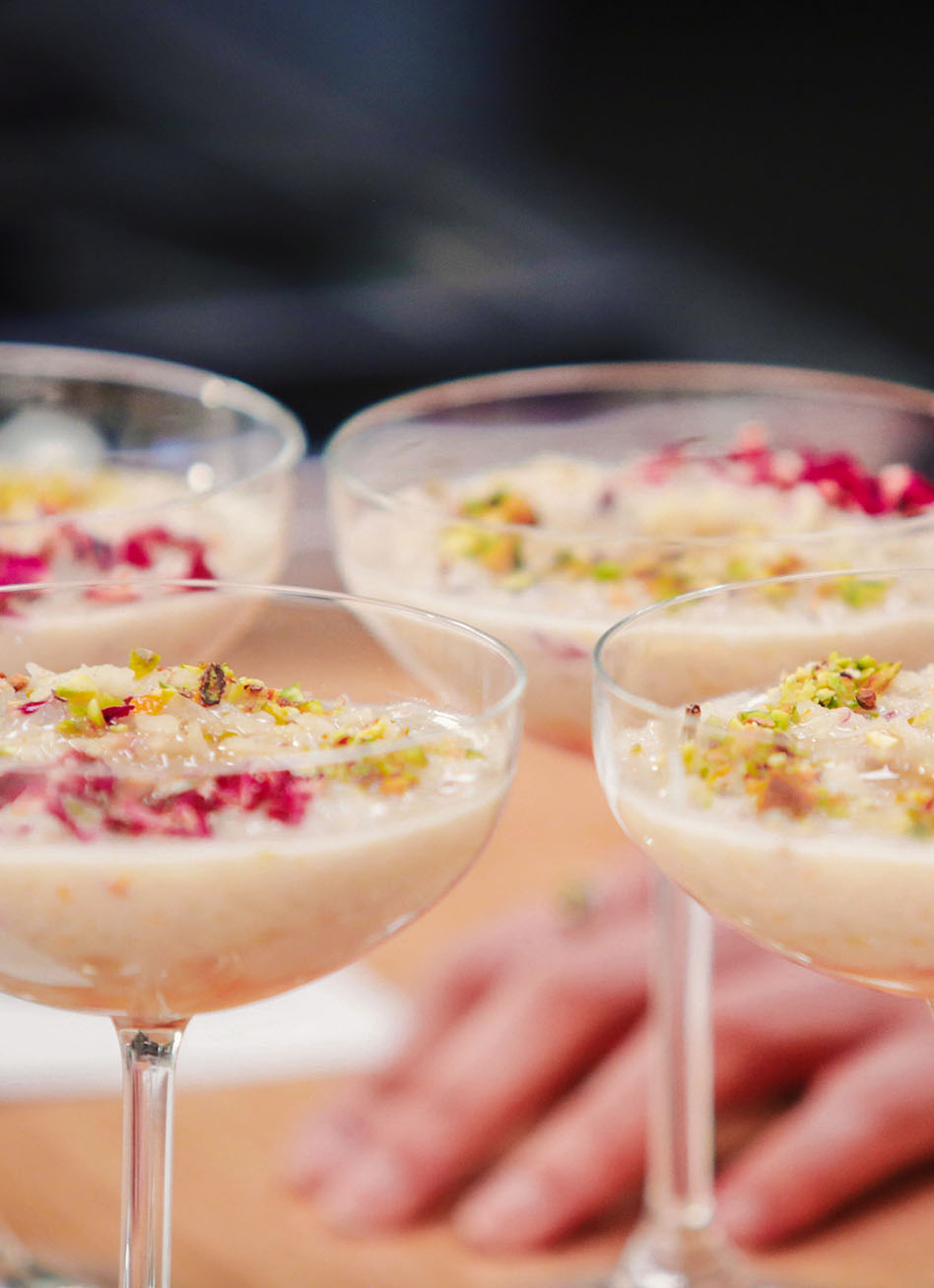 Four cocktail glasses are filled with freshly made rice pudding ornamented with flower pedals and pistachio nuts. The pudding rests on the counter top of the studio kitchen at Particle Studio in Seattle, WA