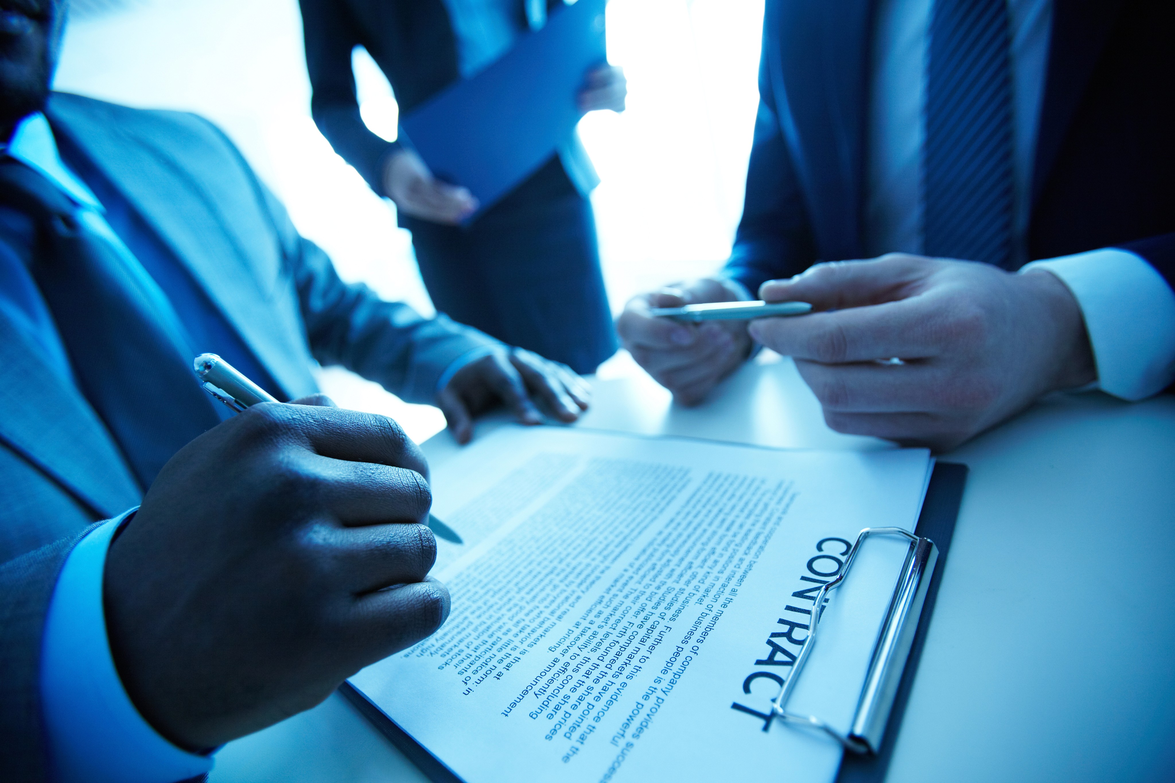 Three individuals in formal business attire are gathered around a table, with two of them signing a contract document, emphasizing a professional business agreement setting.