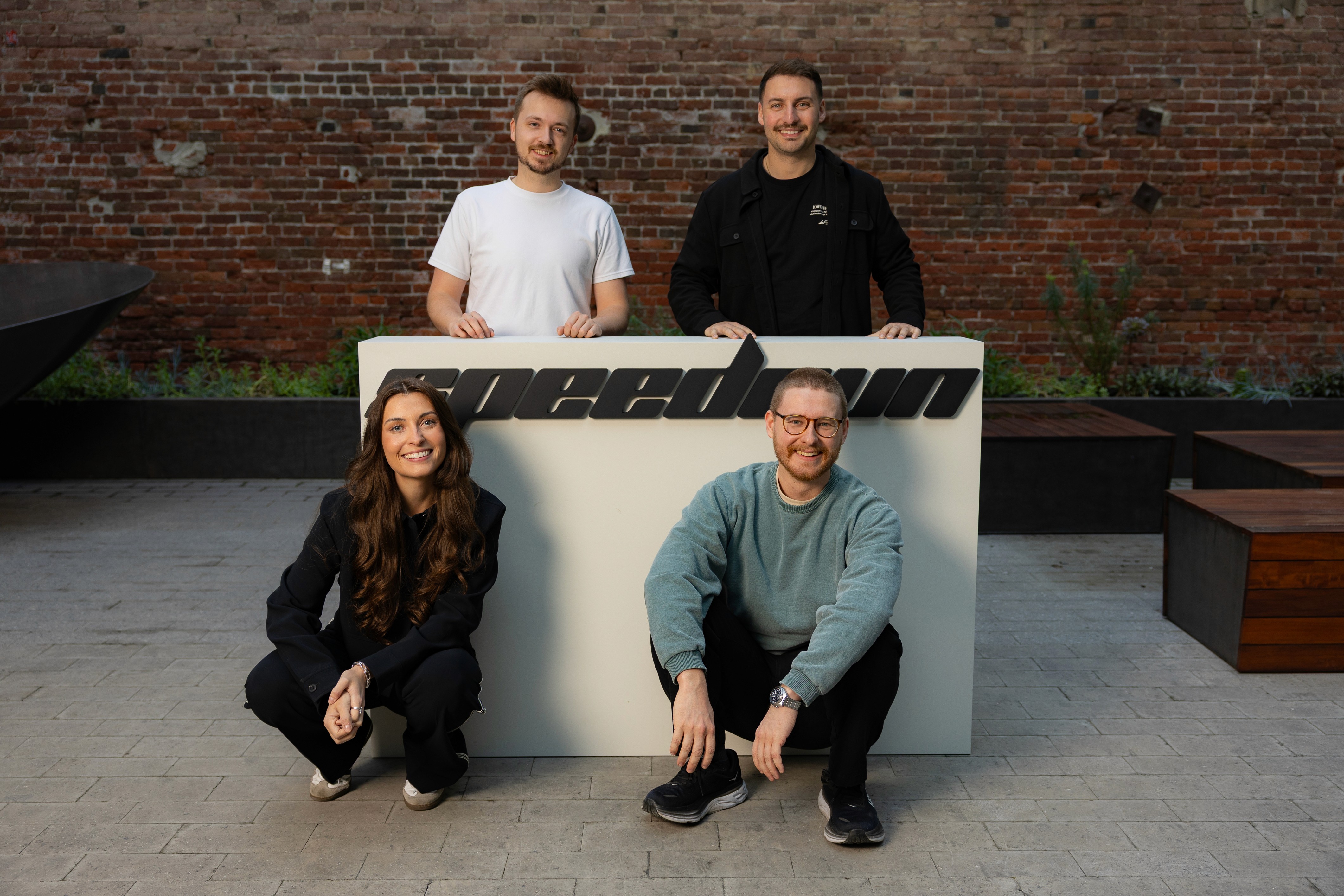 The four founders of Pluvo pose with a large white sign featuring the word "speedrun" against a brick wall background to announce their seed round funding.