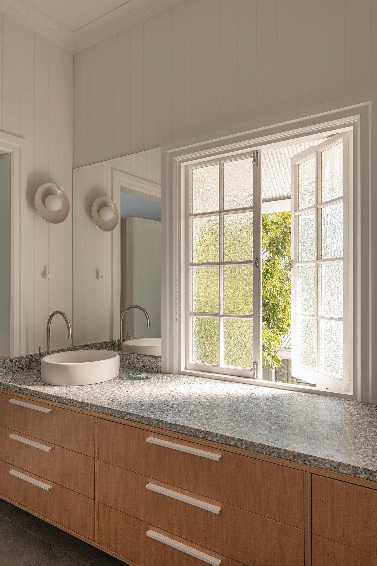 Bathroom vanity with timber joinery, stone benchtop, round basin, and a textured-glass window bringing in soft daylight.