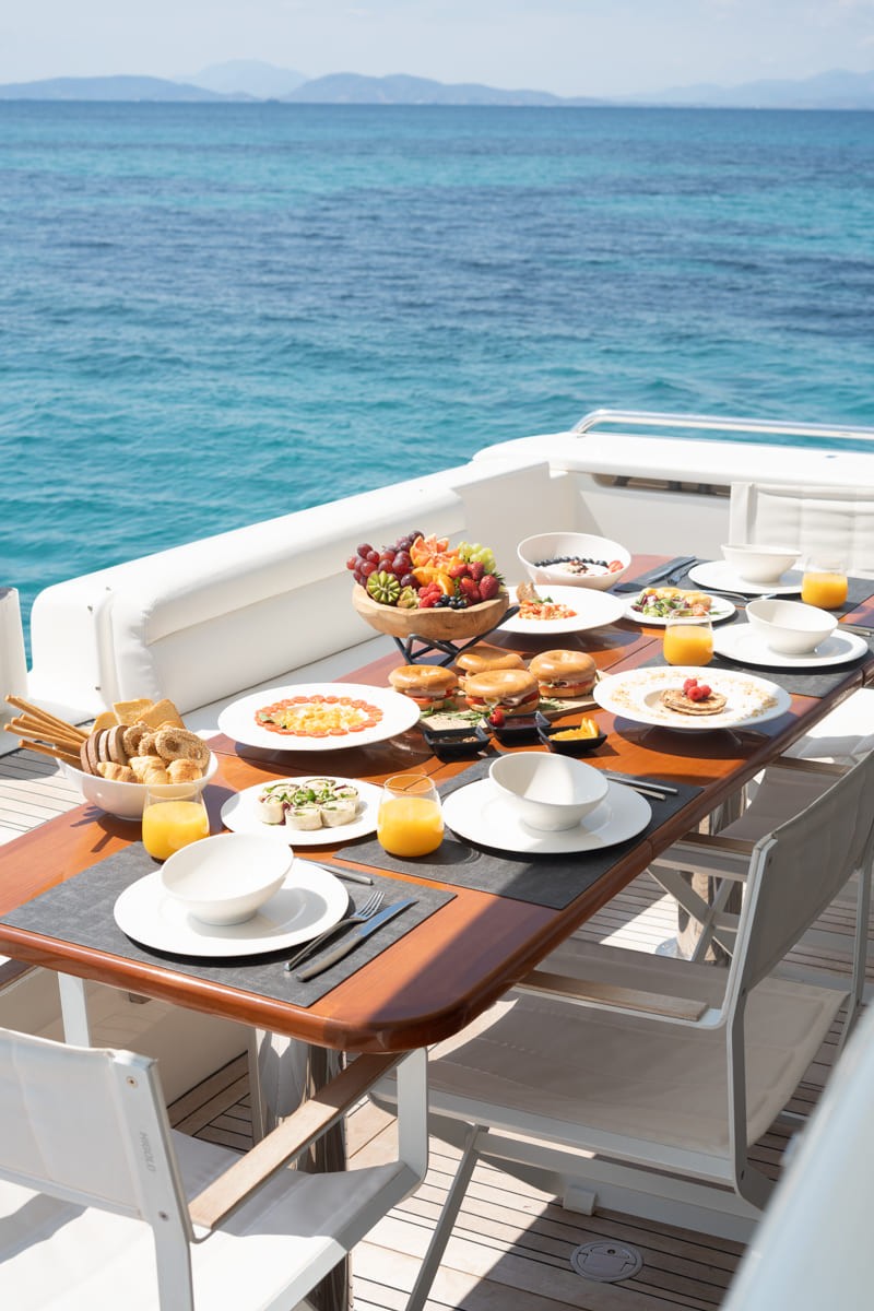 Breakfast table set on yacht deck with fresh fruit, pastries, and orange juice overlooking turquoise Aegean waters and distant Greek islands.