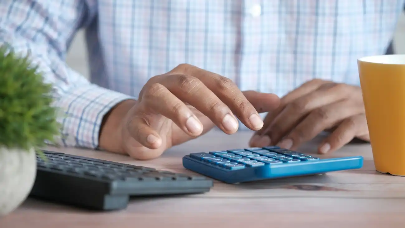Hands using a blue calculator next to a laptop on a desk with a coffee cup — illustrating tax and bookkeeping for club sponsorship, including invoicing and tracking income thresholds.