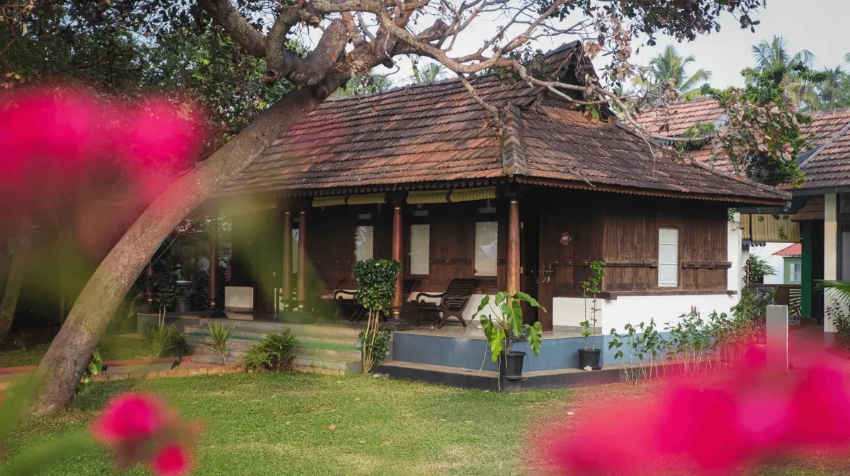 Heritage Room cottage at Vedic Village Kodungallur with carved wooden pillars, a terracotta tiled roof and pink bougainvillea in the foreground