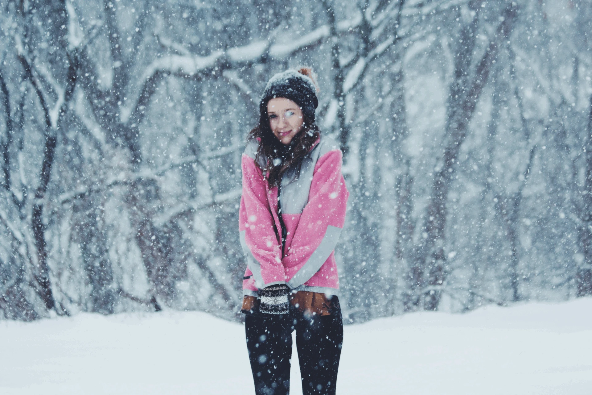 Woman in a pink and gray winter jacket standing in a snowy forest during active snowfall.