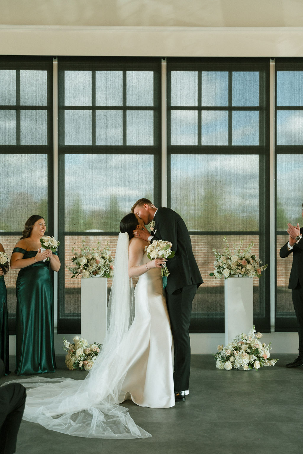 Couple kisses in an indoor wedding venue, white and pink floral arrangements surround them.