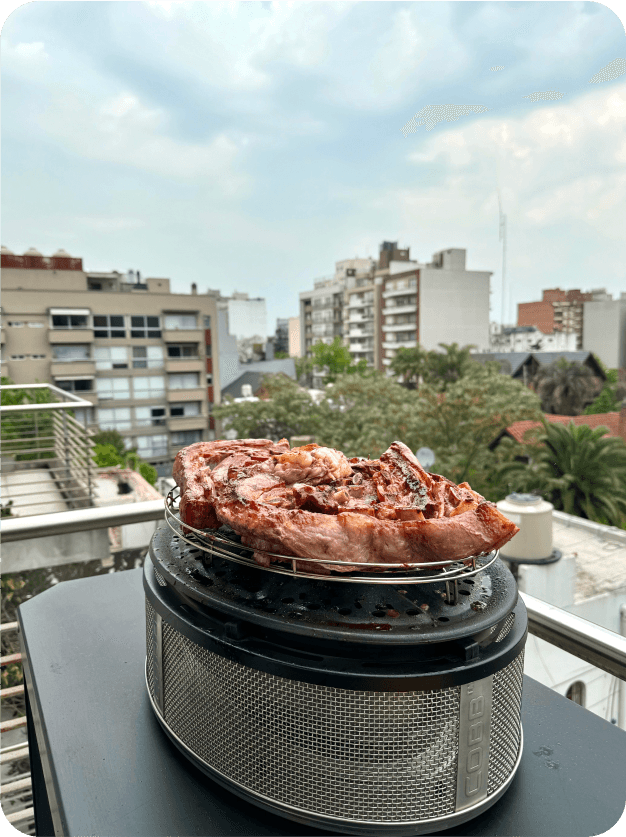Parrilla COBB Supreme con carne asandose sobre una mesa en el balcón de un departamento