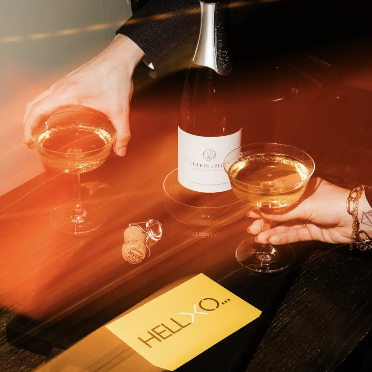 Hands holding coupe glasses beside a bottle of sparkling wine and a HELLO note.