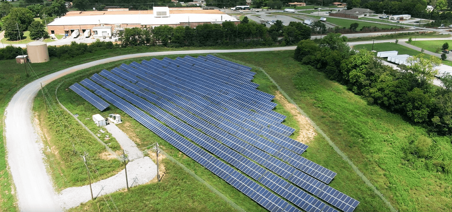 Large solar farm with rows of panels installed on open land near industrial buildings.