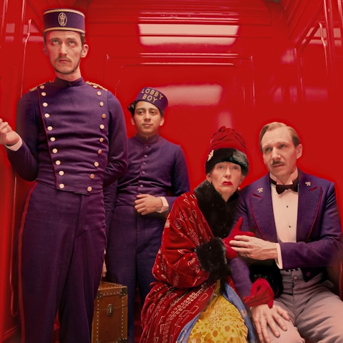 Four people in elaborate period costumes stand and sit in a brightly lit red room, with two dressed as bellhops and two in formal attire.