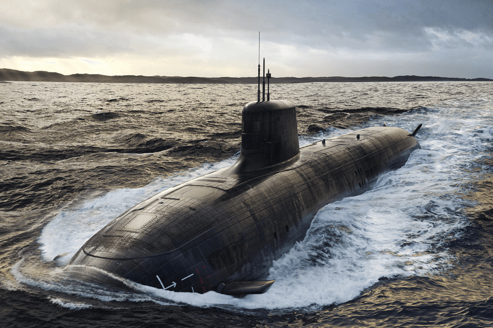 A large black submarine travels on the surface of the sea under a cloudy sky, with distant land visible on the horizon.