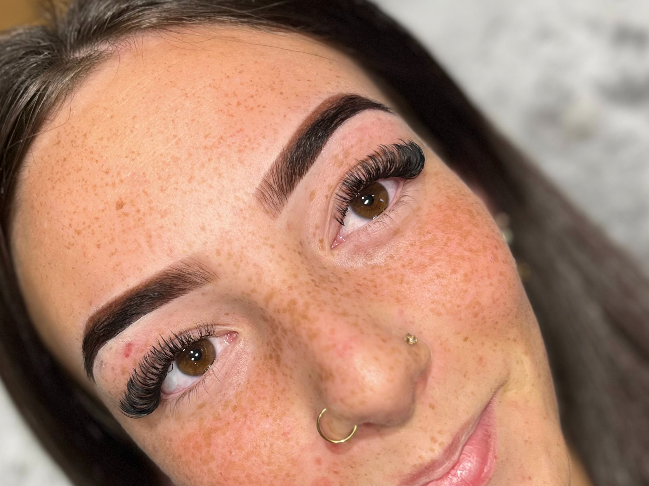 Close-up of a woman's face showing flawless makeup with bold eyebrows, long lashes, and a nose piercing. Her skin is freckled, conveying a natural look.