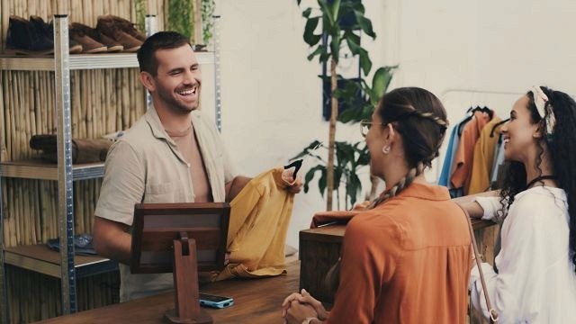 Homem branco atrás do balcão, segurando cabide com peça de roupa, sorri para duas mulheres em um ambiente de loja