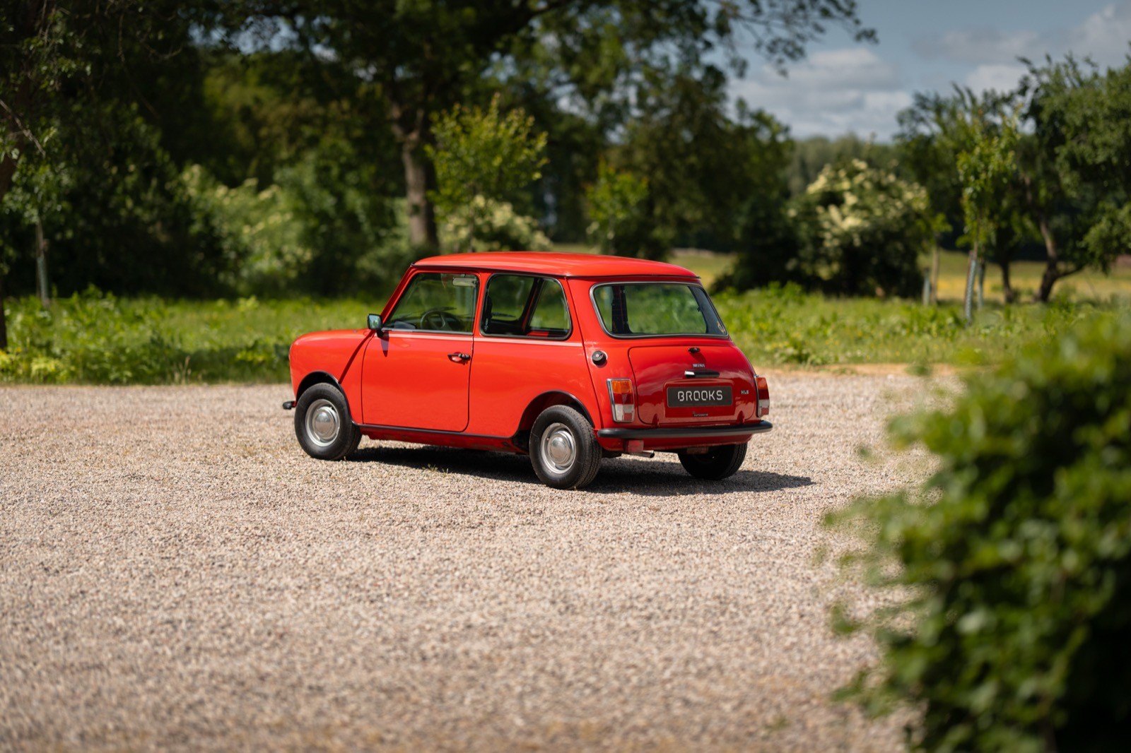 A red vintage car parked on a gravel driveway surrounded by green trees and a clear blue sky.