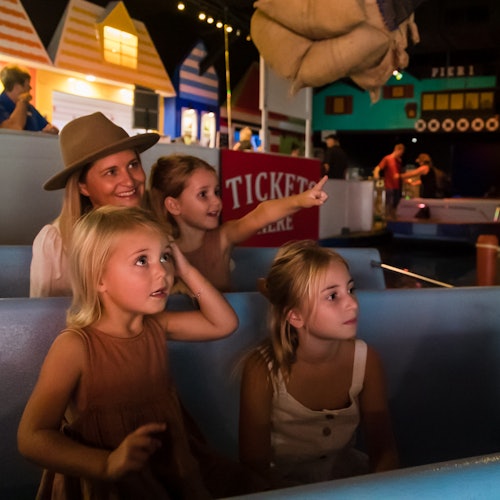 A woman and three children are seated in a ride, looking excitedly at something overhead. Colorful buildings are in the background.