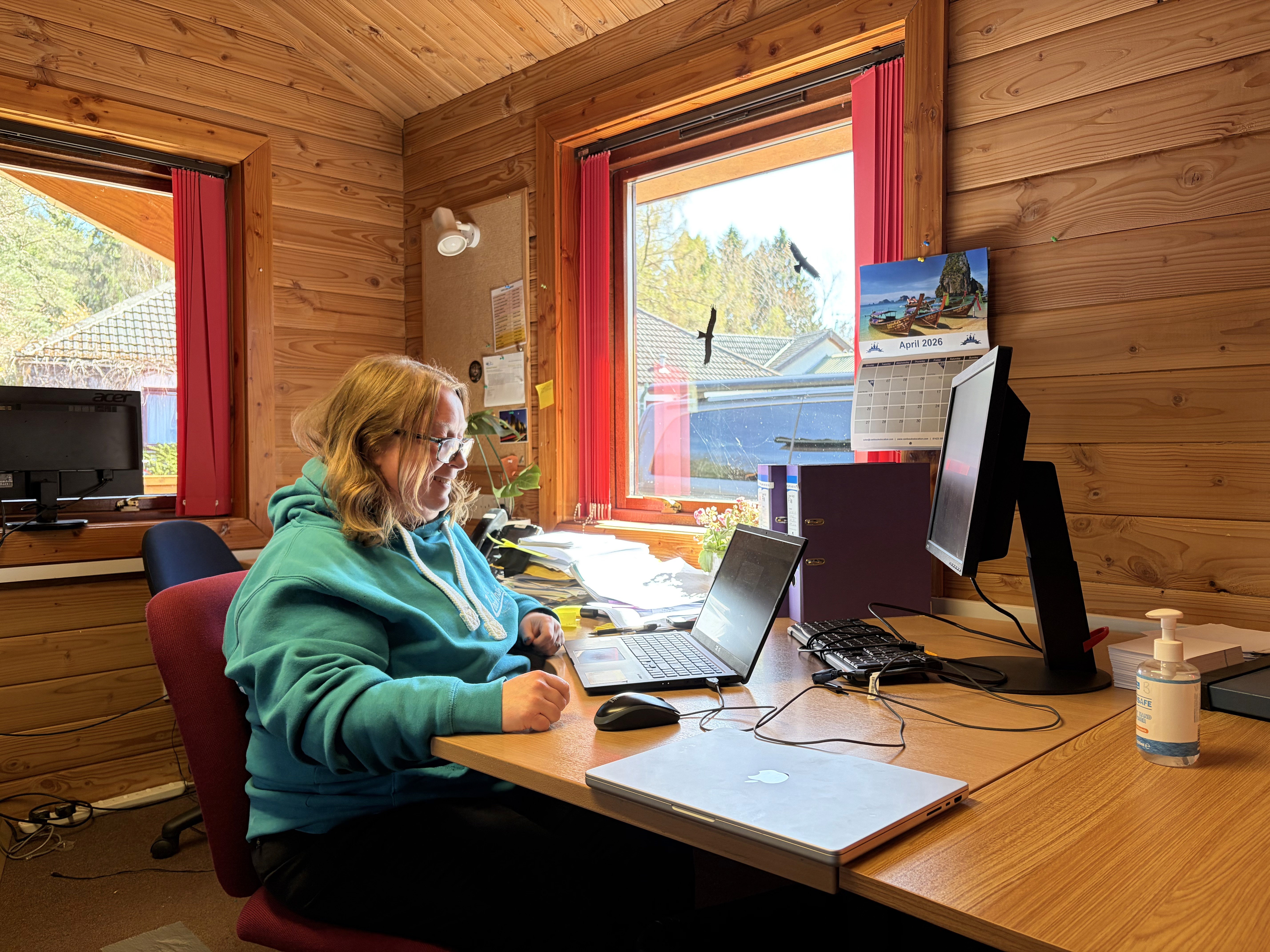 A small boat-shaped money box labeled "RNLI Money Box" is on a wooden surface beside a card payment terminal. The background shows a cozy, wood-paneled room.