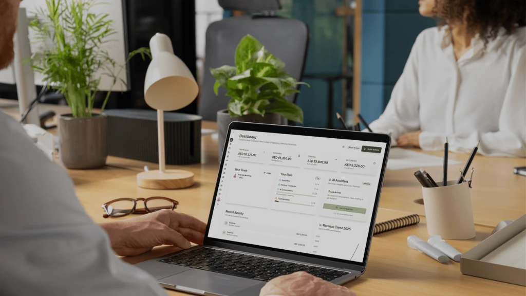 A person's hand is shown working on a laptop displaying charts, while another person is seated nearby in a cozy setting.