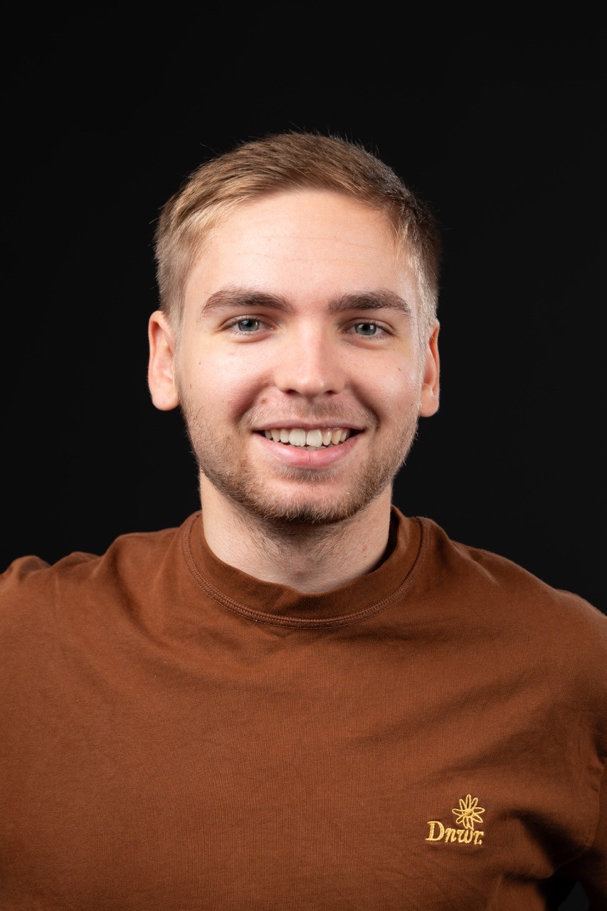 Portrait studio d'Adrien Delebecq, homme blond aux yeux bleu avec un t-shirt marron, sur fond noir.