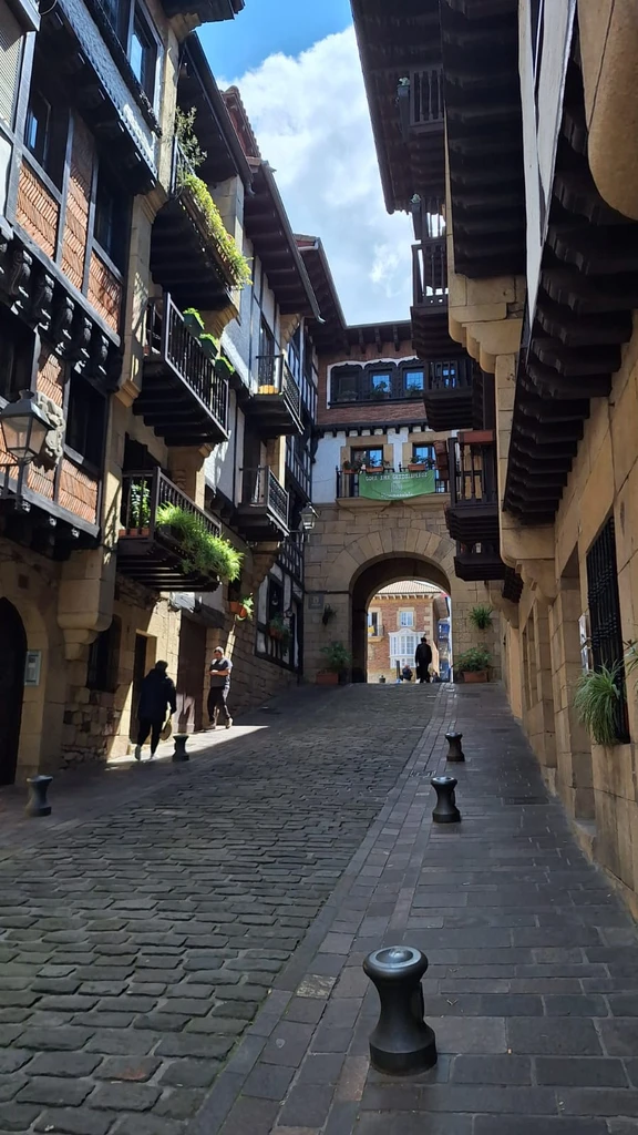 Narrow cobblestone street in a historic town, buildings with balconies on either side, archway at the end of the street.