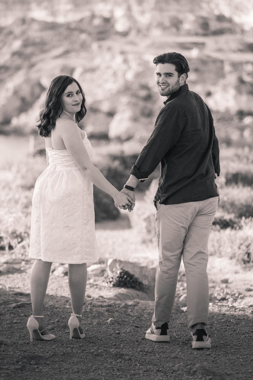 A groom in a dark suit carries a bride in a white dress across a sunny, grassy field. The couple shares a joyful, intimate moment with trees in the background.