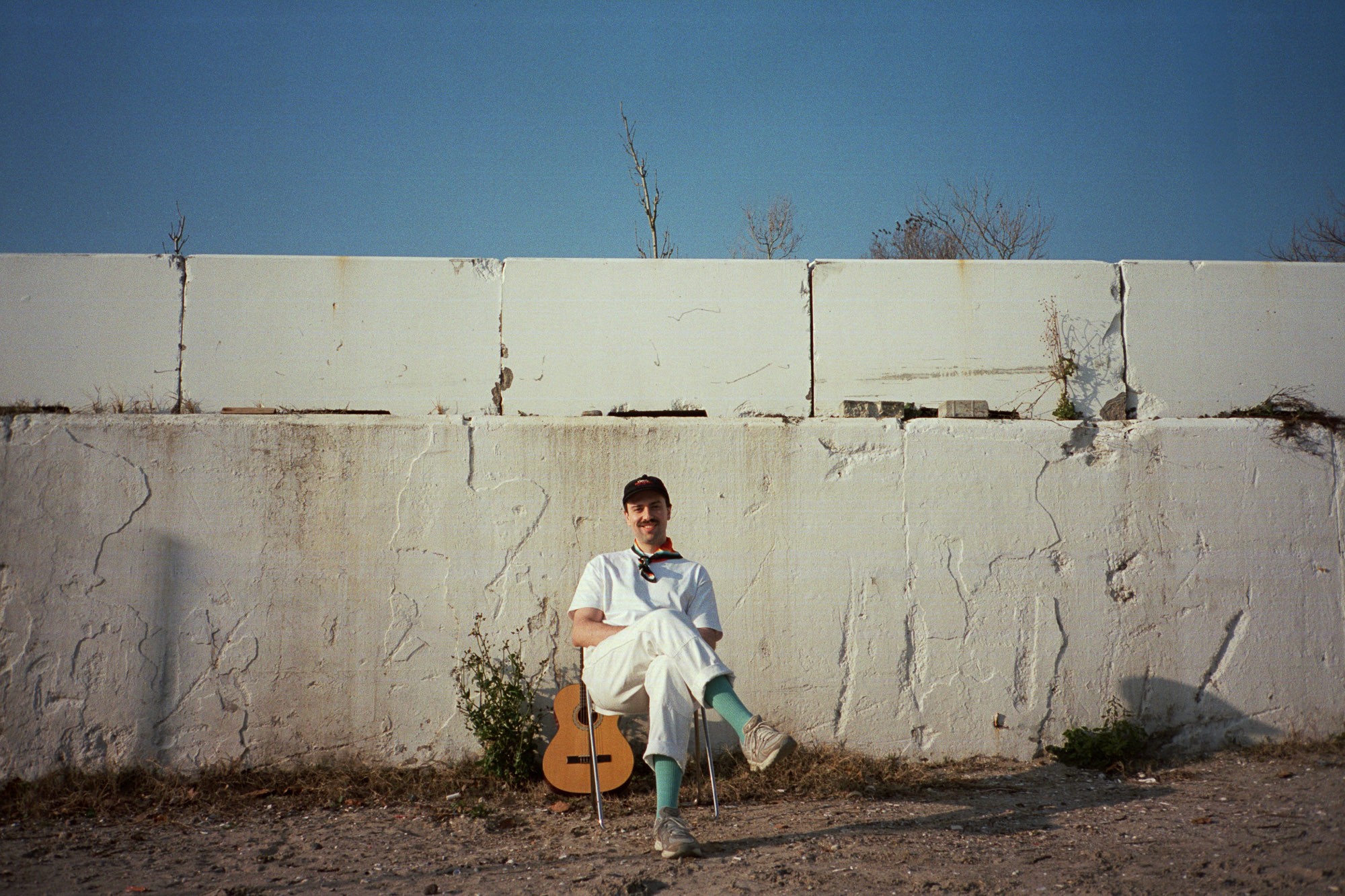 A person sits on a chair against a large, weathered white concrete wall, with a guitar propped beside them, under a clear blue sky.