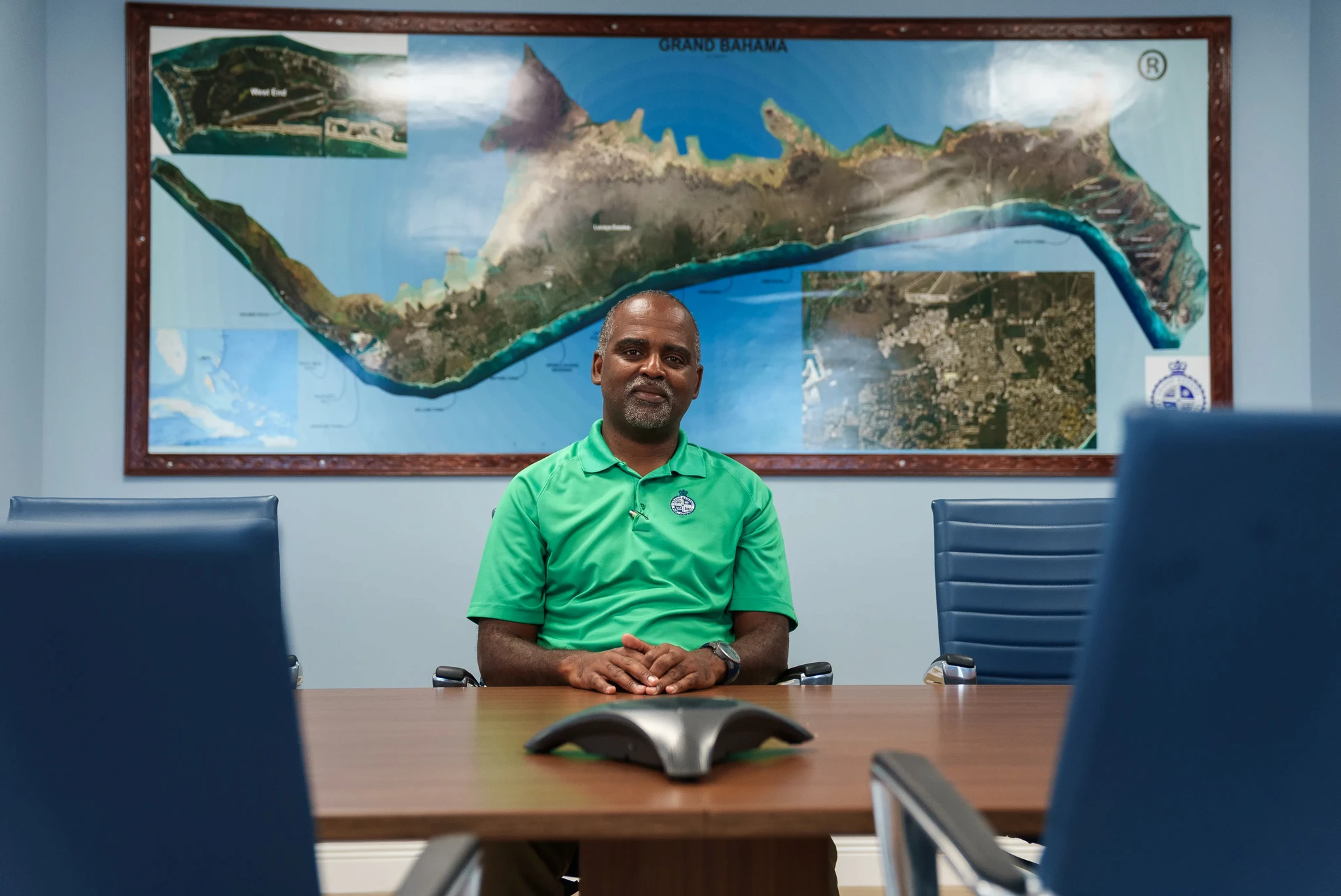 A person in a green shirt sits at a conference table with blue chairs. A large map of Grand Bahama Island is on the wall behind them.