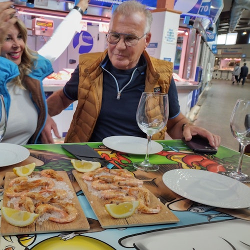 Two people sit at a table with plates of shrimp and lemon wedges, empty wine glasses, and colorful cartoon table mat.
