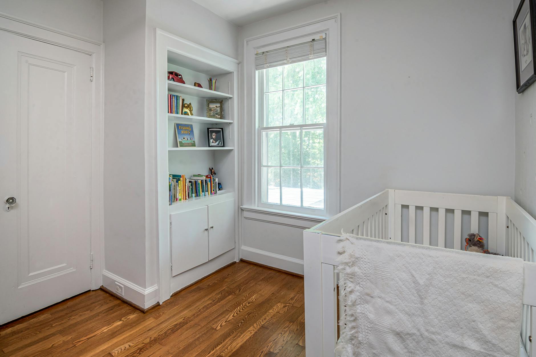 A bright room with a large window, light gray walls, wooden floor, and a white crib against the wall.