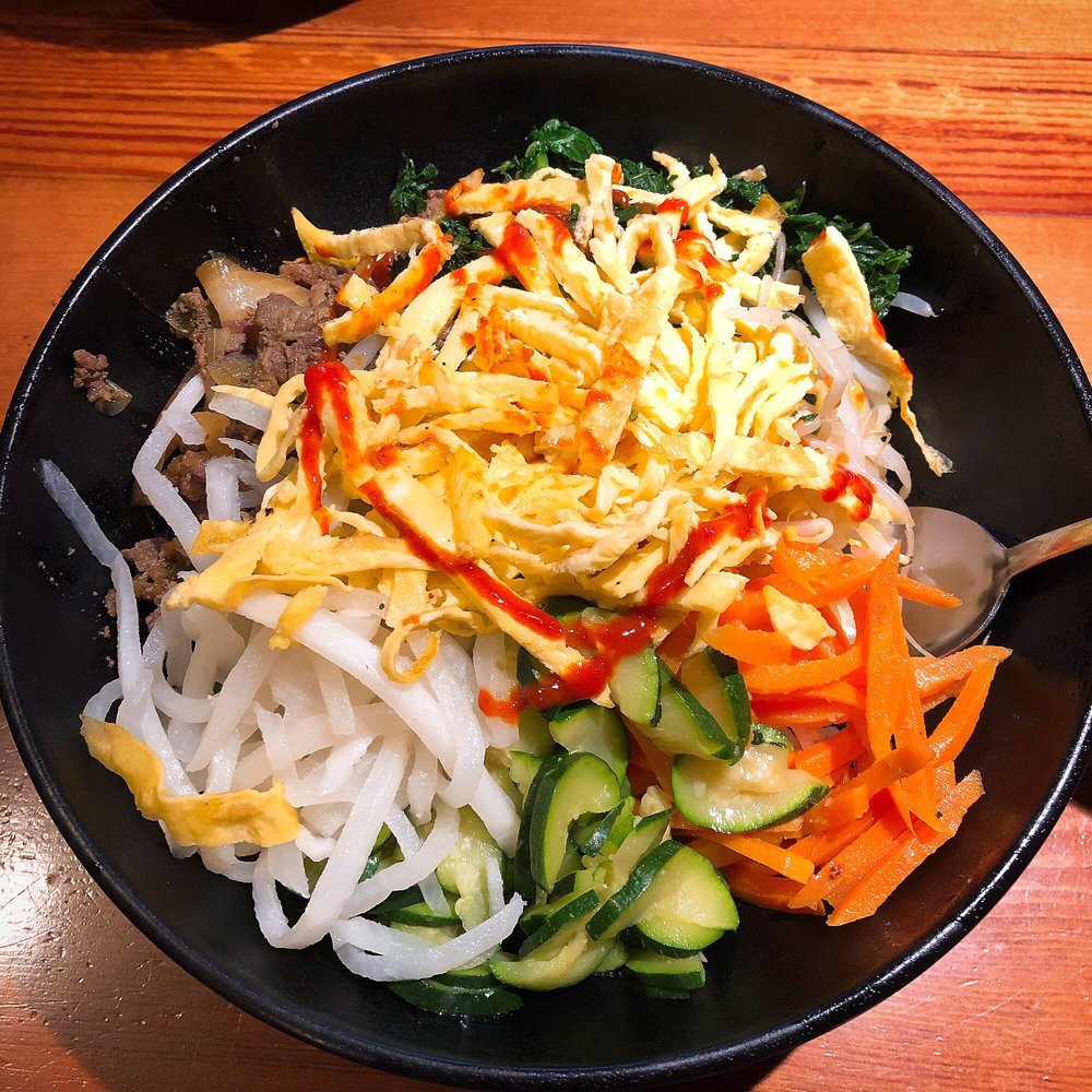 A colorful bowl of Bibimbap featuring perfectly cooked rice, fresh vegetables, seasoned beef, and a sunny-side-up egg drizzled with gochujang sauce. A staple Korean comfort dish at K-Bap & Wings.